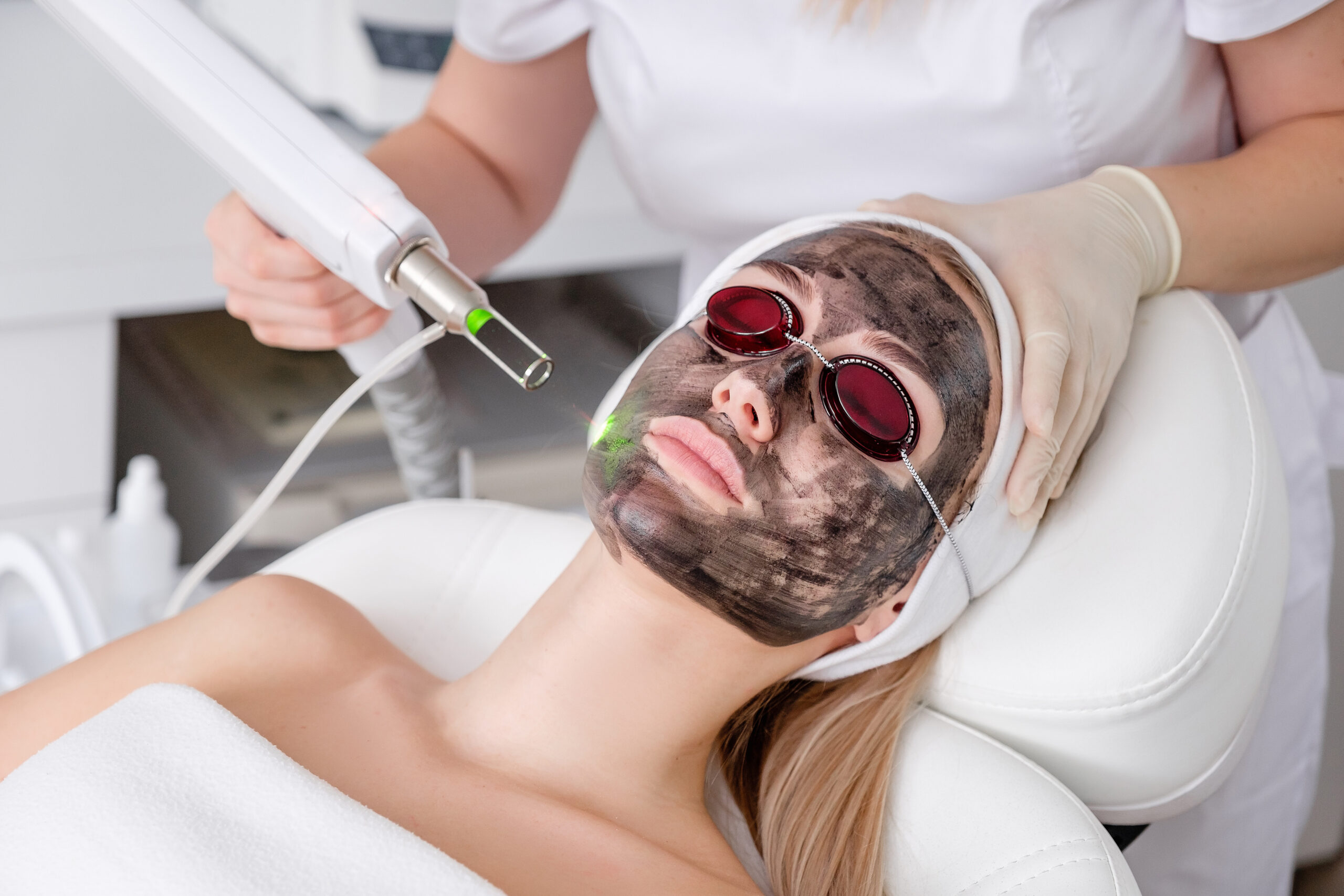 Close-up of cosmetologist's hand making hardware carbon peeling by young pretty customer woman in safety glasses headdress lying on couch in beauty salon. Facial cleaning concept. Cosmetology Services