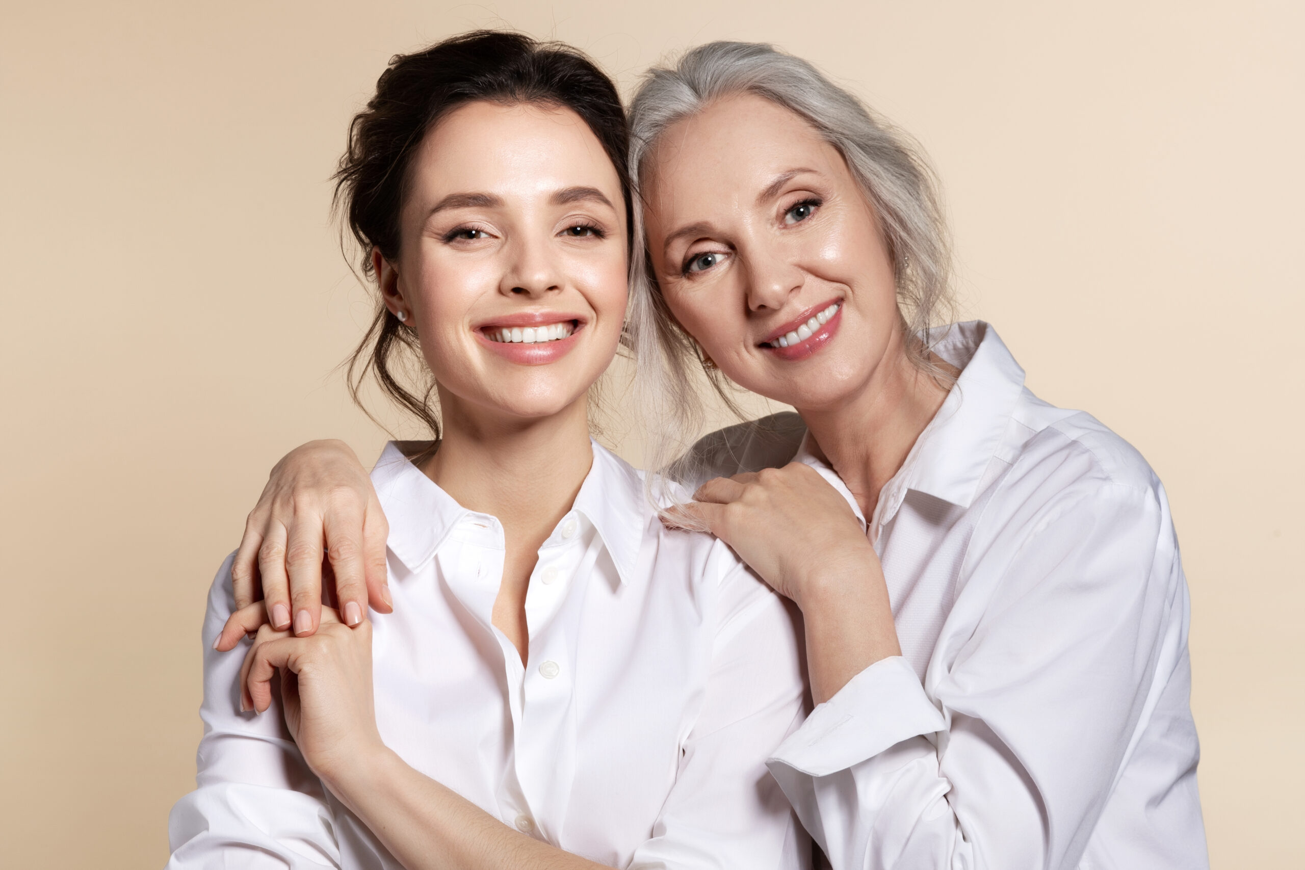 Two beautiful woman in white shirt hug face forward portrait. Studio headshot of toothy smiling grandmother and granddaughter, senior mother and daughter, elderly and young sister. Happy family