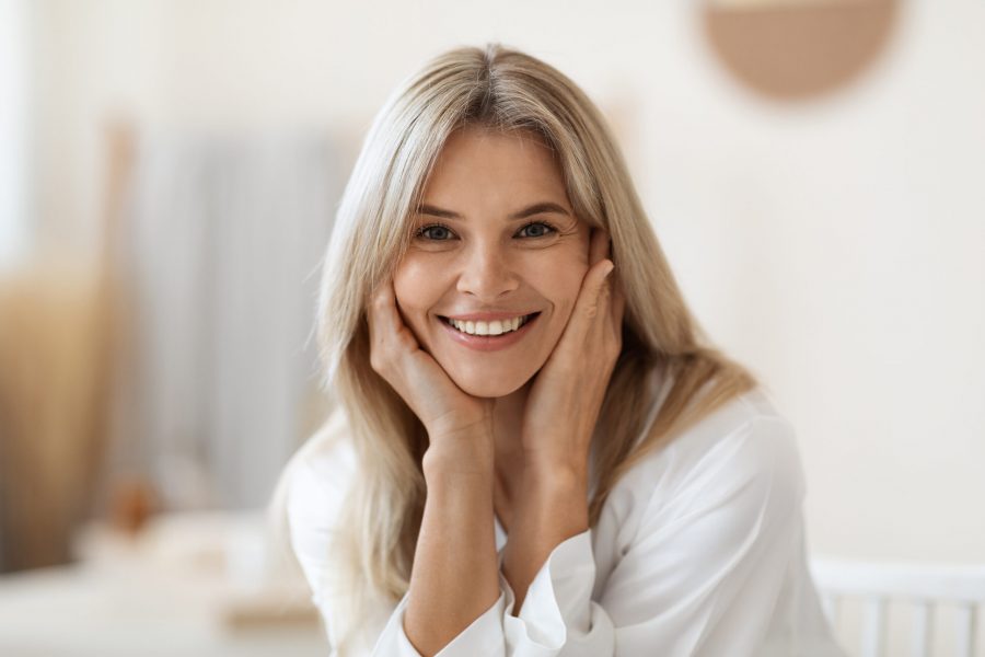 Beautiful blonde middle aged woman smiling at camera, touching her face, attractive lady doing morning face care routine, white bathroom interior, blurred background, closeup portrait, copy space