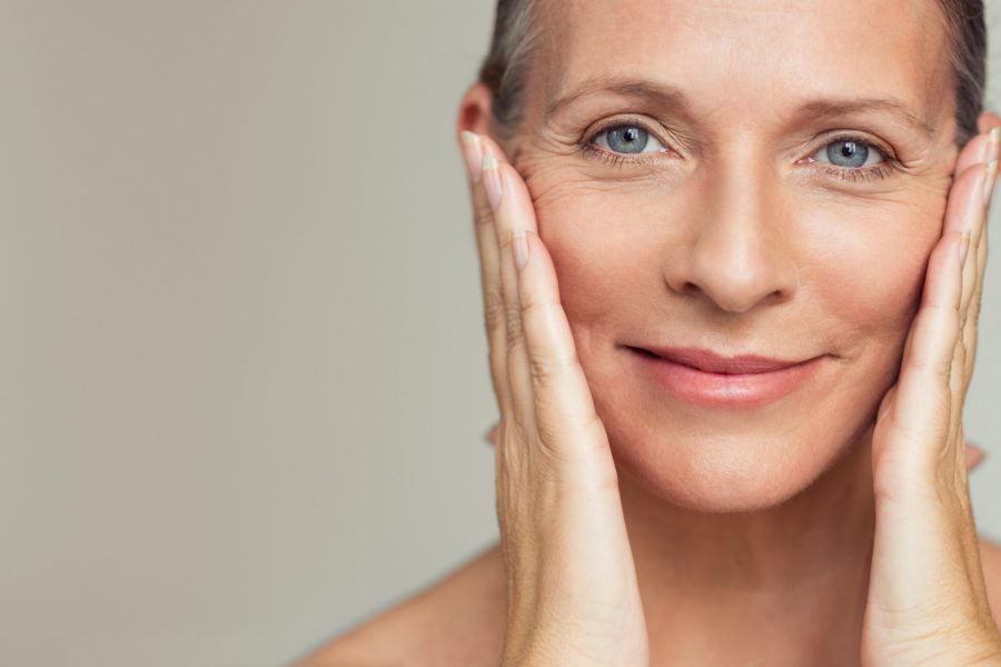Portrait of beautiful senior woman touching her perfect skin and looking at camera. Closeup face of mature woman with wrinkles massaging face isolated over grey background. Aging process concept.